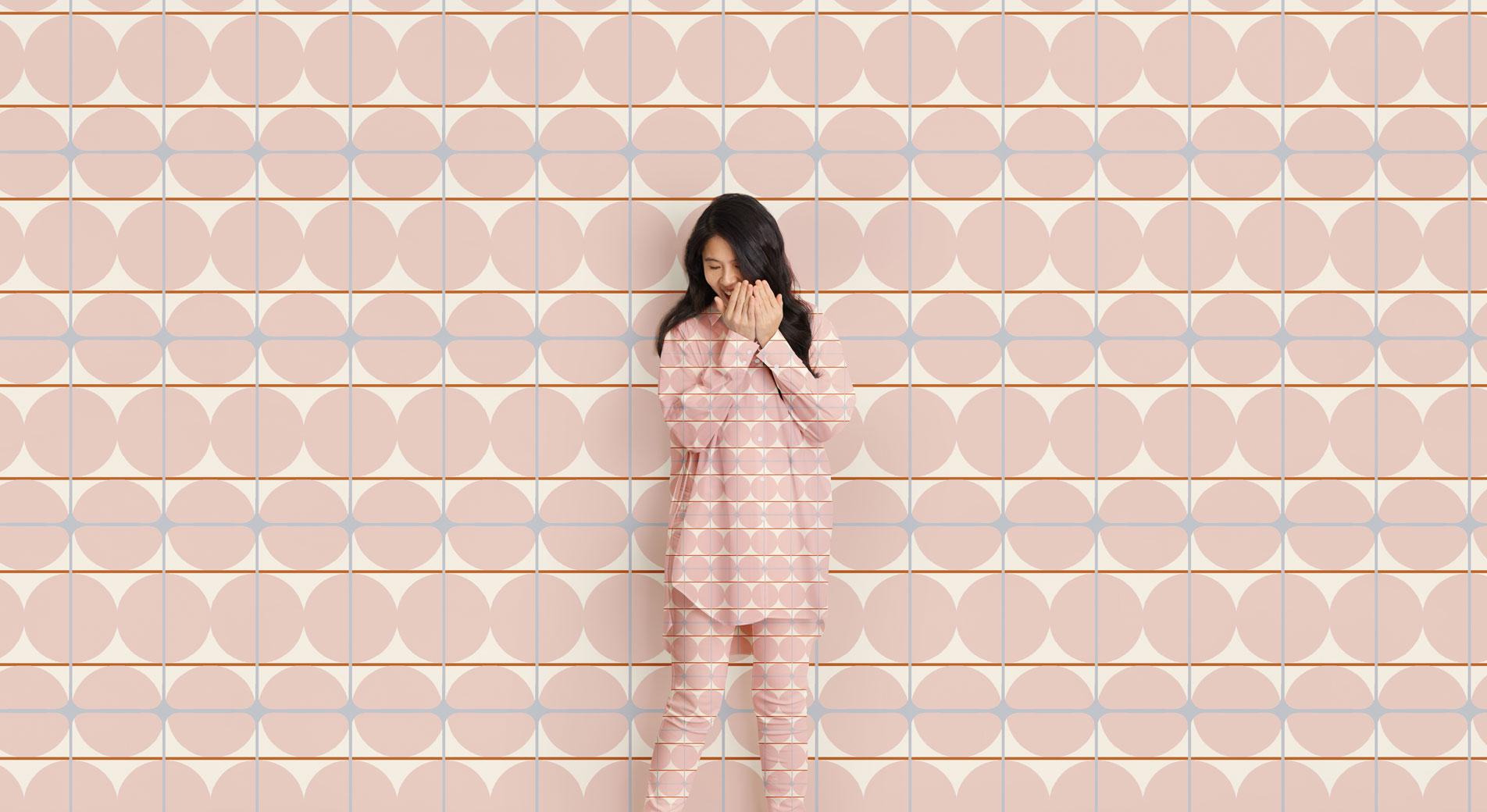 A woman stands, giggling and covering her face in front of a geometric print background. Her shirt and pants are in the same matching pattern.